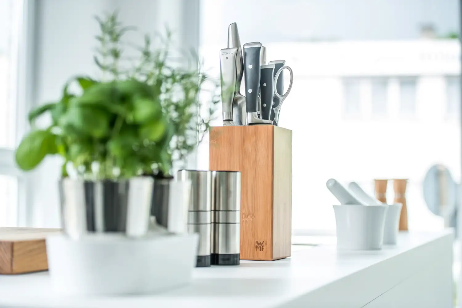 Küchenmesser und Schere in einem Holzblock neben frischen Kräutern in Töpfen und Gewürzmühlen auf weißer Küchenarbeitsplatte. Im Hintergrund stehen noch zwei weiße Mörser.