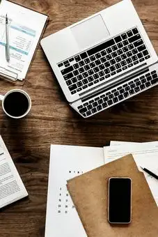 Auf einem Holztisch liegen ein aufgeklappter Laptop, ein Smartphone auf einem braunen Umschlag, mehrere Dokumente, zwei Stifte und eine Tasse Kaffee.