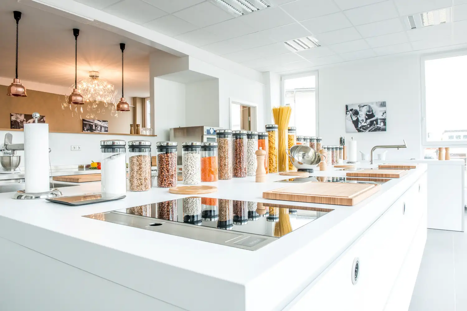 Moderne, helle Küche mit weißen Arbeitsflächen, mehreren Induktionskochfeldern und einer Reihe von transparenten Vorratsgläsern mit verschiedenen Hülsenfrüchten und Nudeln.