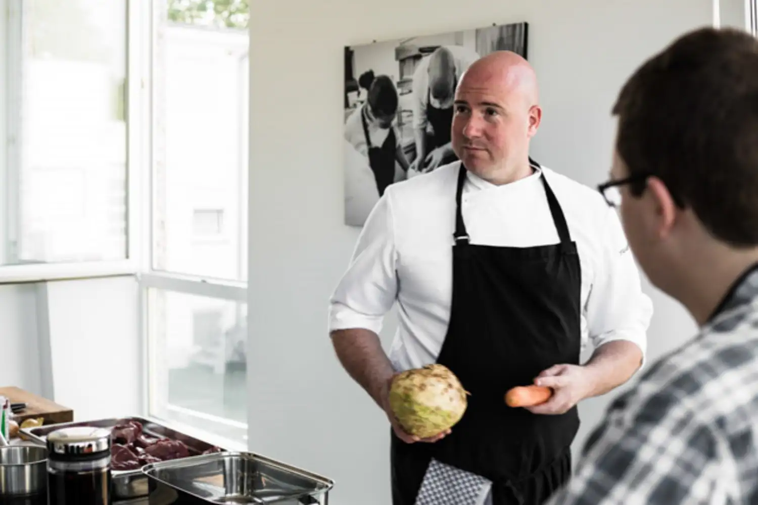 Koch in weißer Jacke und schwarzer Schürze hält ein Sellerieknolle und eine Karotte und spricht mit einer Person.