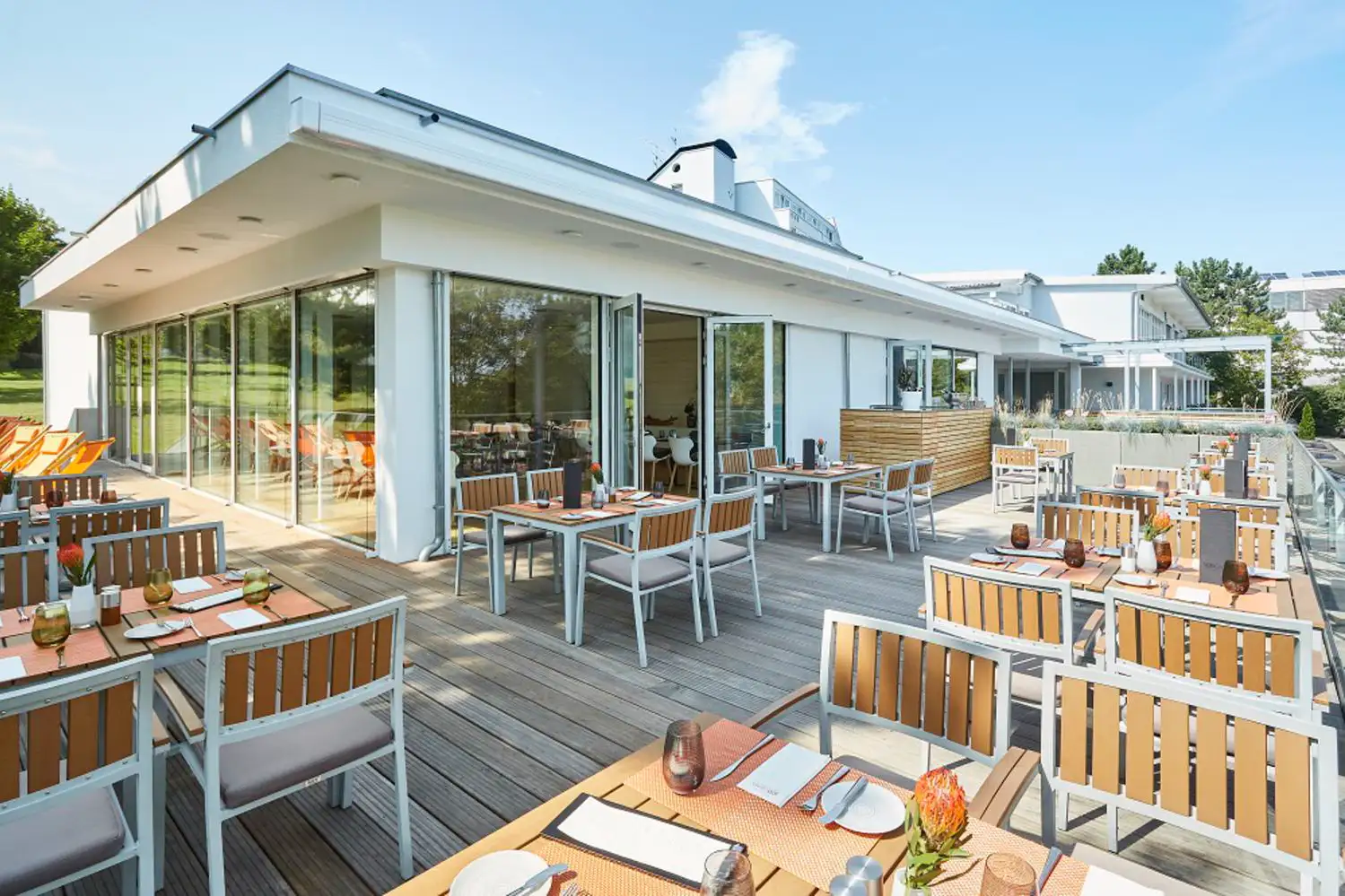 Moderner Außenbereich eines Restaurants mit Holzterrasse, gedeckten Tischen und Stühlen unter klarem Himmel.