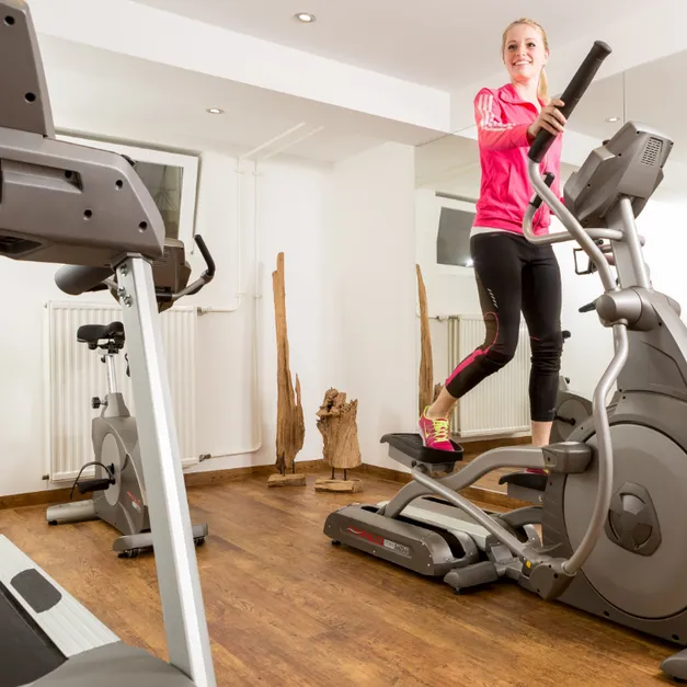 Frau in pinker Sportjacke trainiert auf einem Crosstrainer in einem hellen Fitnessraum mit Holzboden.