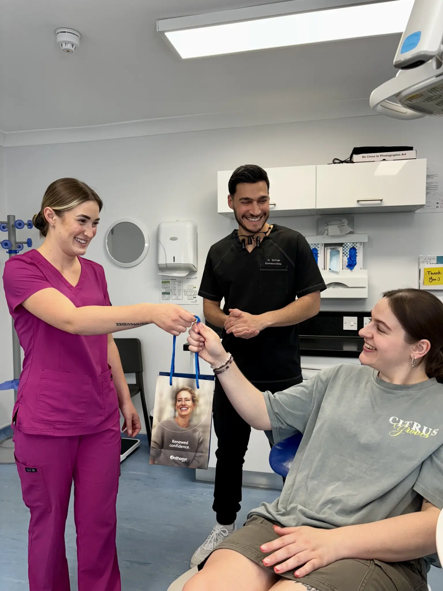 A female nurse in purple scrubs hands a gift bag to a smiling young woman seated in a dental chair, while a male dentist in black scrubs watches them smiling.