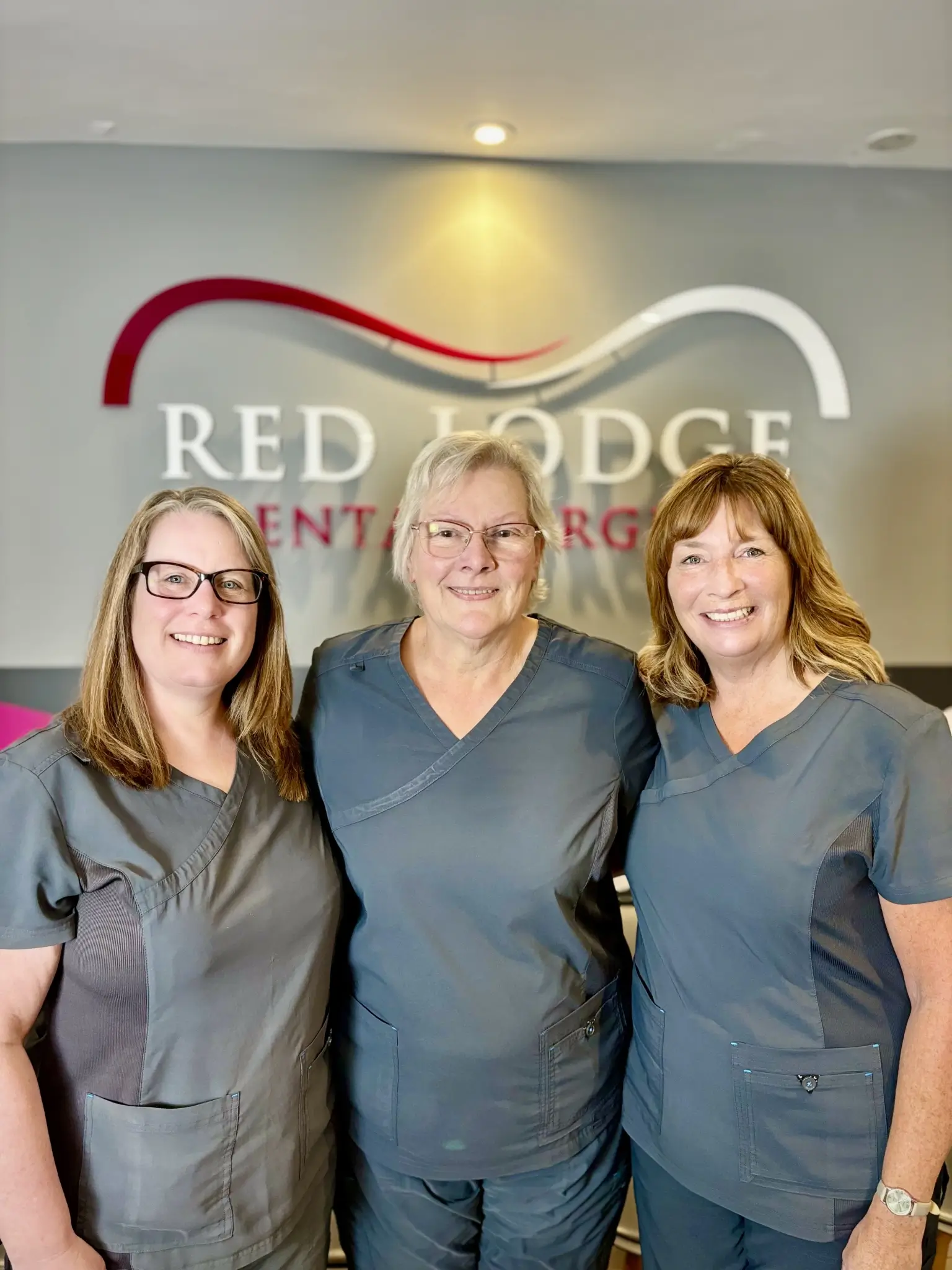Three women in gray medical scrubs smiling, standing together in front of a wall with the Red Lodge Dental Surgery logo.