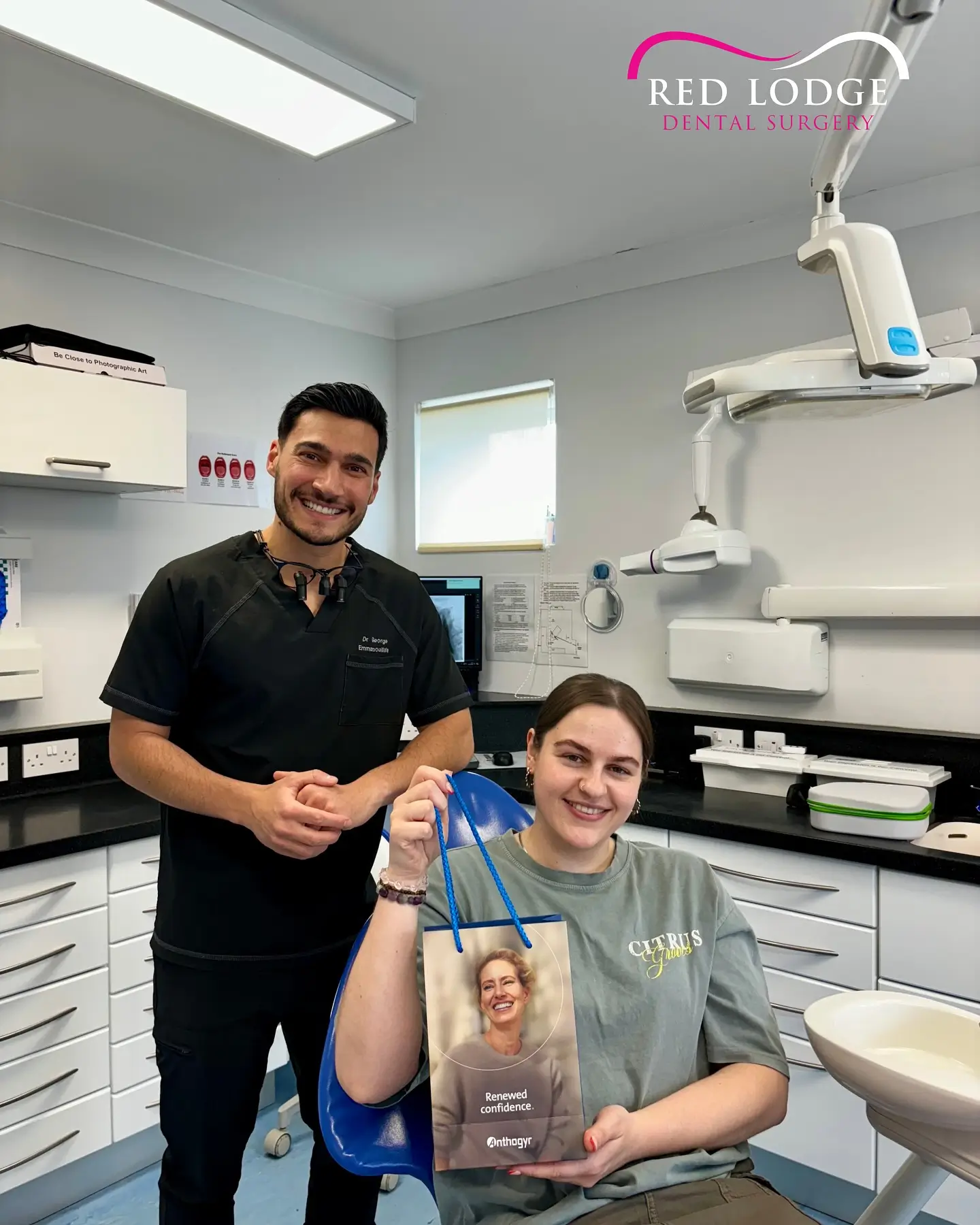 Smiling female patient seated in dental chair holding a gift bag, accompanied by a smiling male dentist in a modern dental clinic.