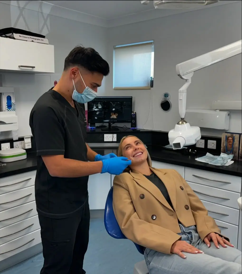 Dentist wearing a face mask and blue gloves examining a smiling patient's mouth in a dental clinic.