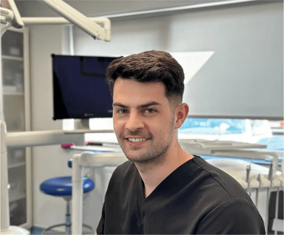 Smiling male dentist wearing black scrubs in a dental office with equipment in the background.