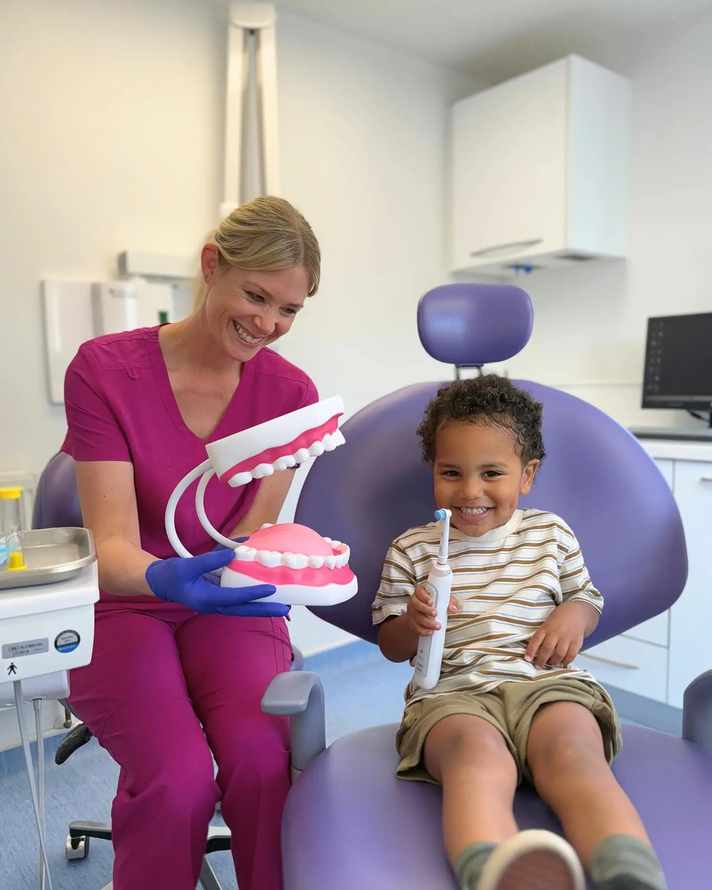 Smiling dentist in purple scrubs showing a large dental model to a happy young boy holding an electric toothbrush in a dental office.