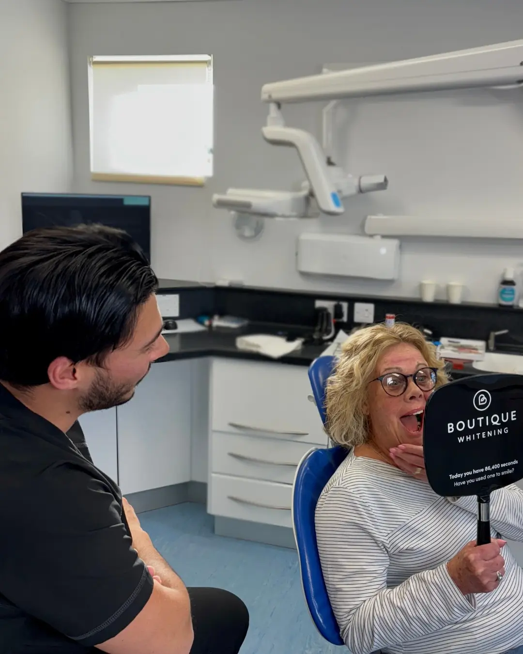 Woman with glasses sitting in a dental chair holding a Boutique Whitening mirror, smiling widely while a male dentist watches her.