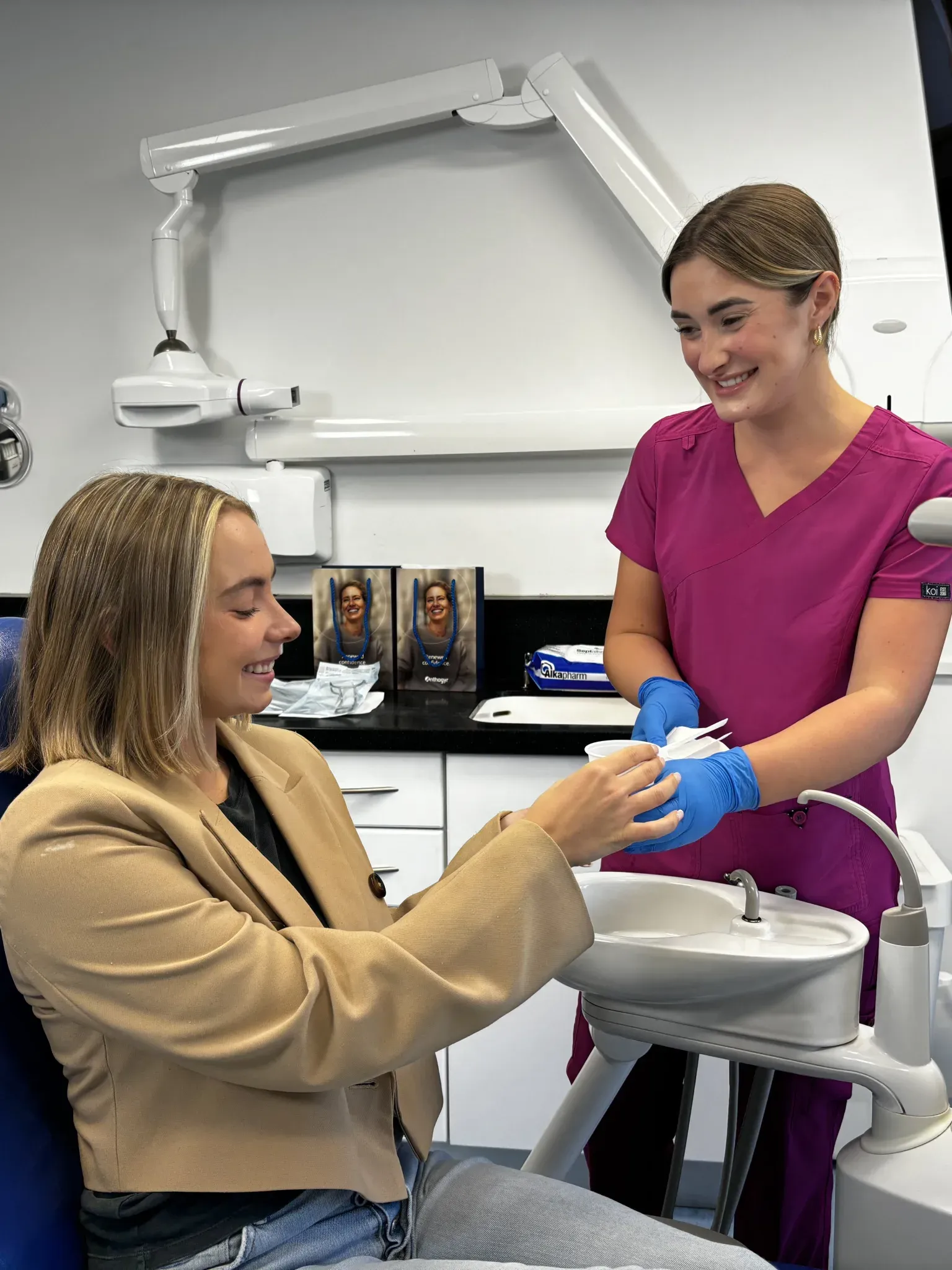 Dental assistant in pink scrubs smiling and handing a dental cup to a seated female patient in a beige jacket.