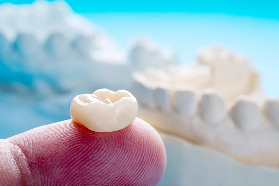 Close-up of a dental crown resting on the tip of a finger with a blurred dental mold in the background.