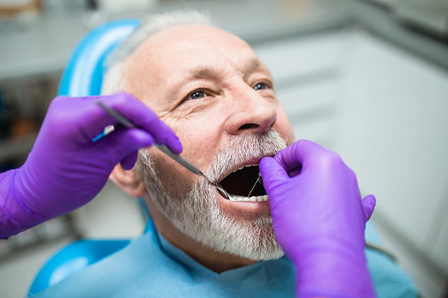 Elderly man with white beard having dental examination by dentist wearing purple gloves in dental clinic.