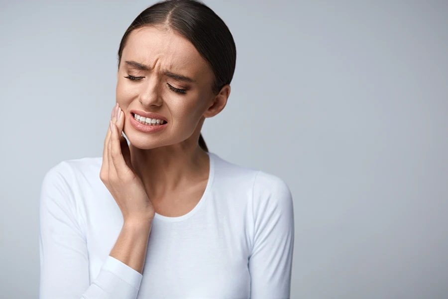 Woman in white shirt holding her cheek with a pained expression indicating toothache or jaw pain.