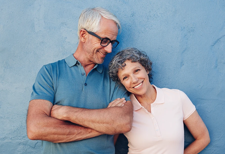 Smiling elderly couple standing close together against a blue wall, woman leaning her head on man's shoulder.