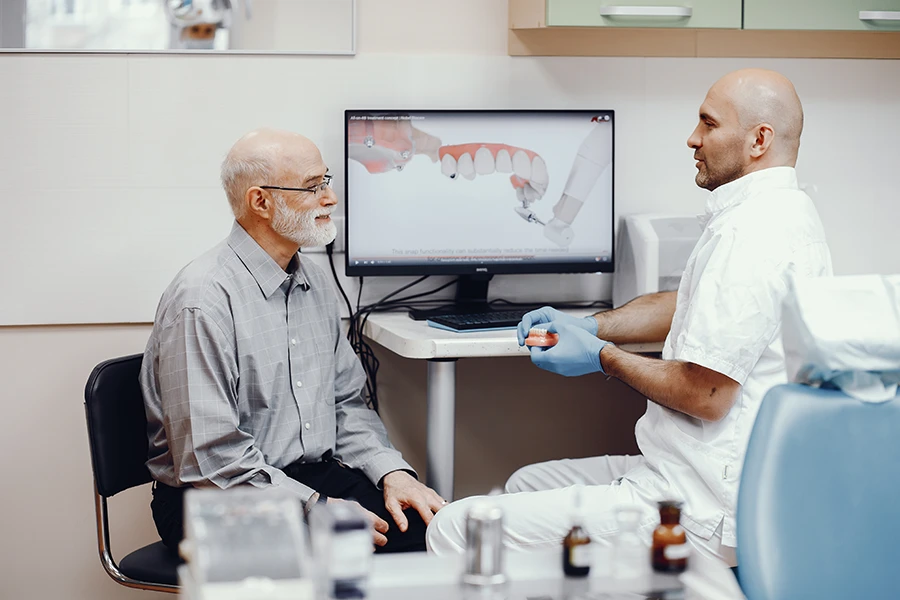 Dentist wearing gloves explaining denture fitting to an elderly man in a dental office with denture images on a computer screen.