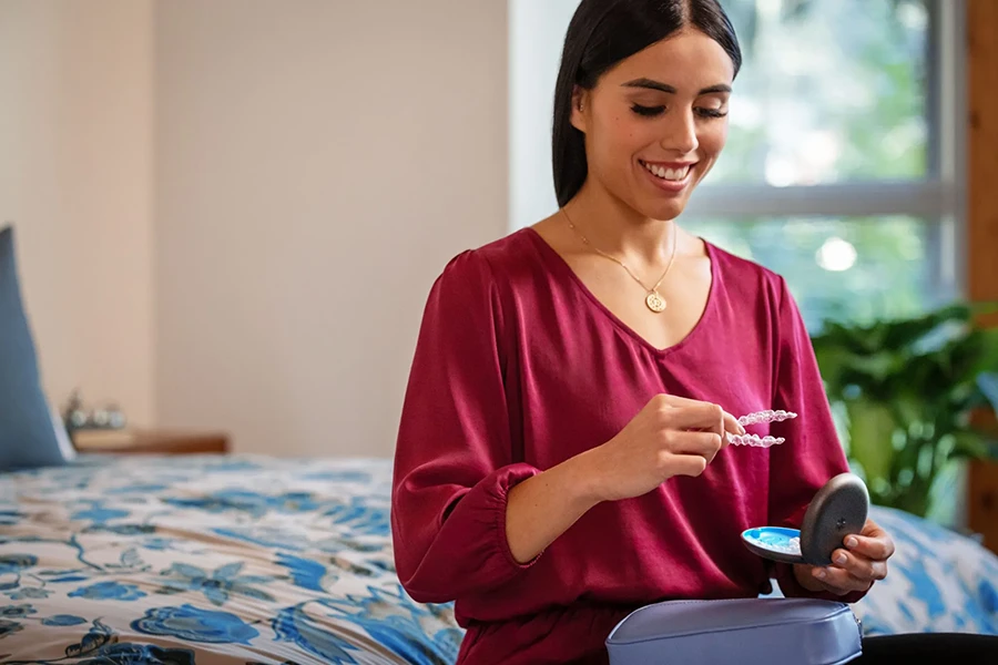 Smiling woman in a red blouse holding a clear dental aligner and its storage case while sitting on a bed.