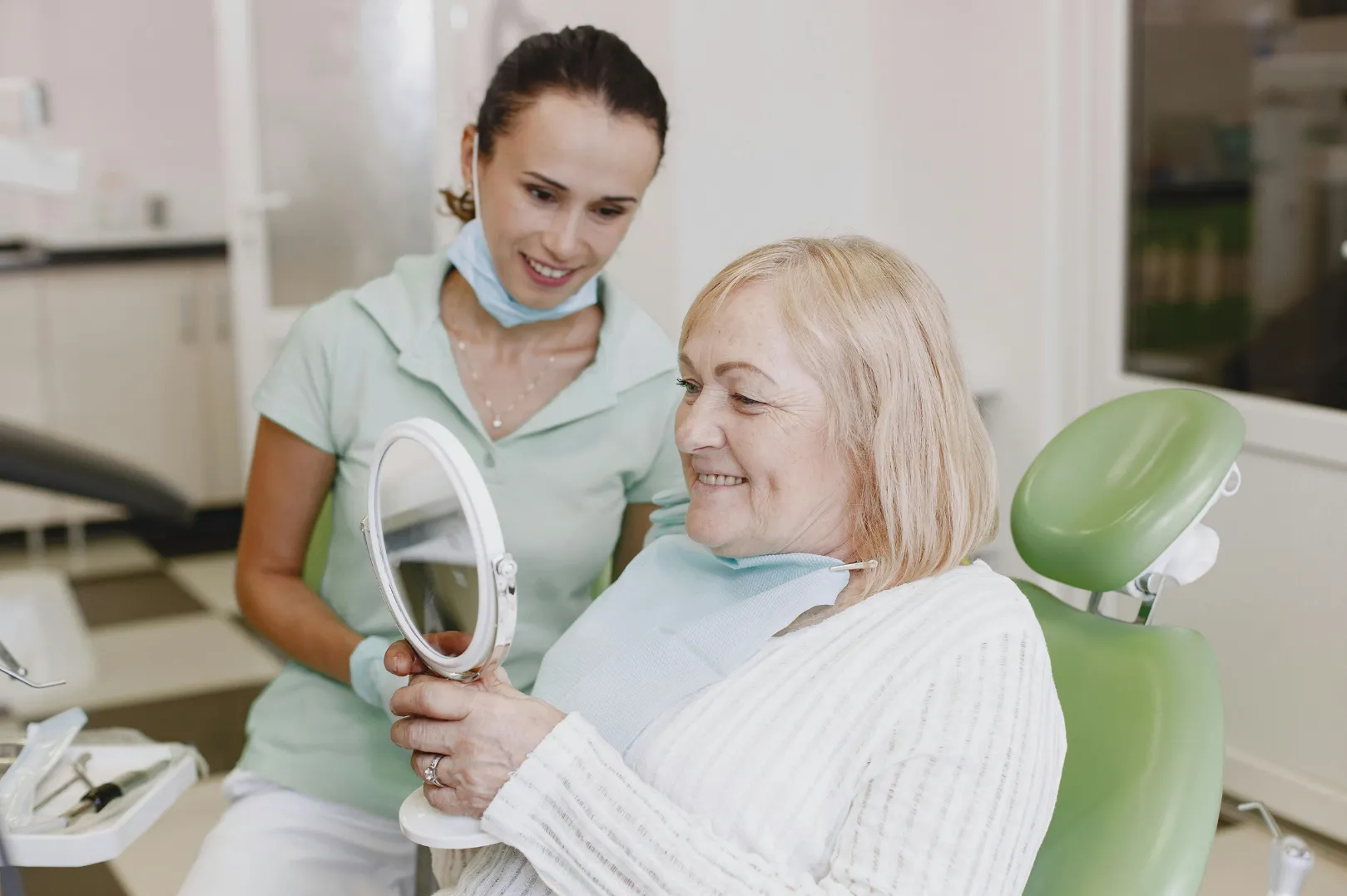 Smiling elderly woman in dental chair holding a round mirror while a dental professional stands behind her.