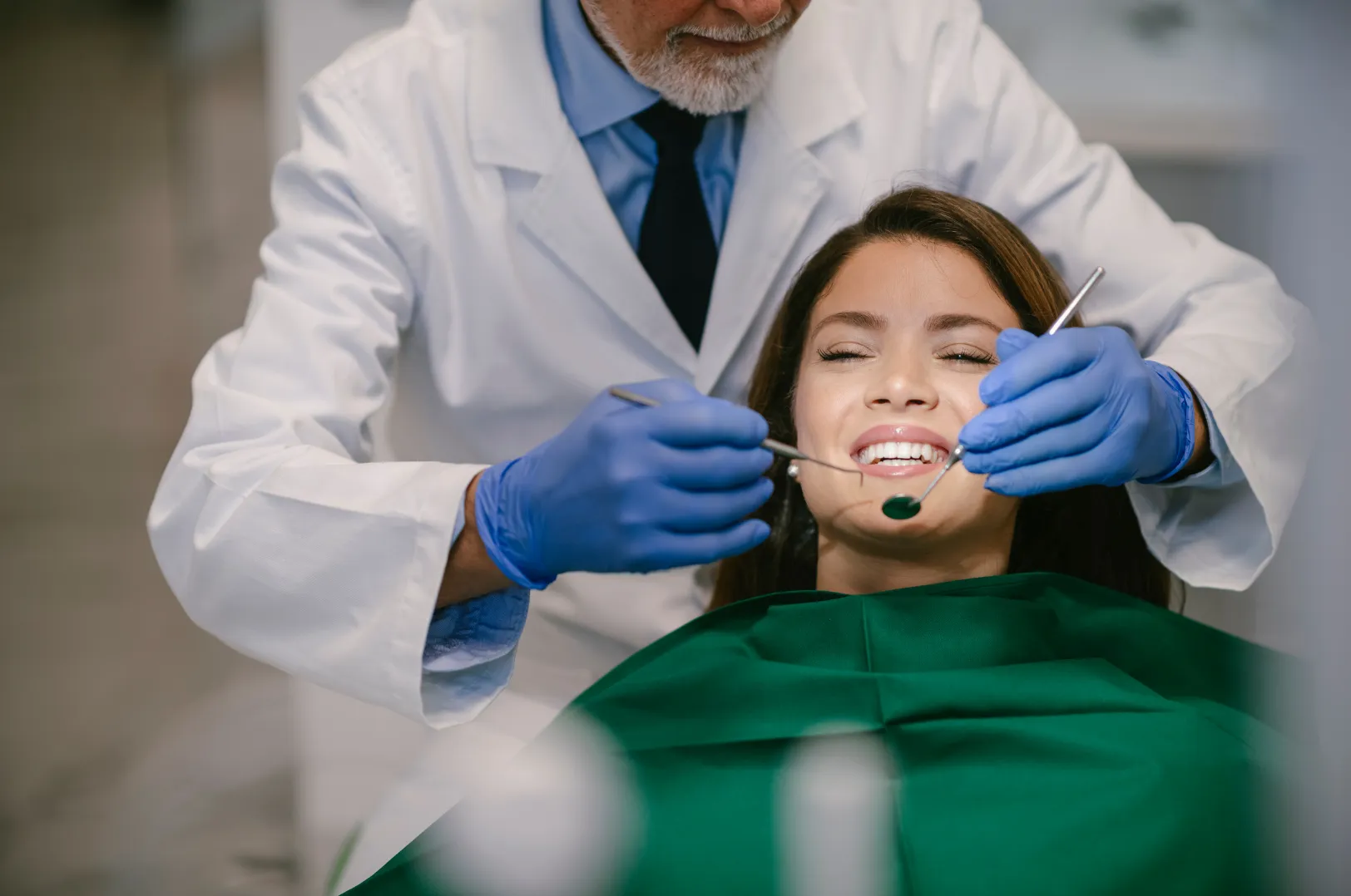 Dentist in white coat and blue gloves examining a smiling female patient's teeth with dental tools.