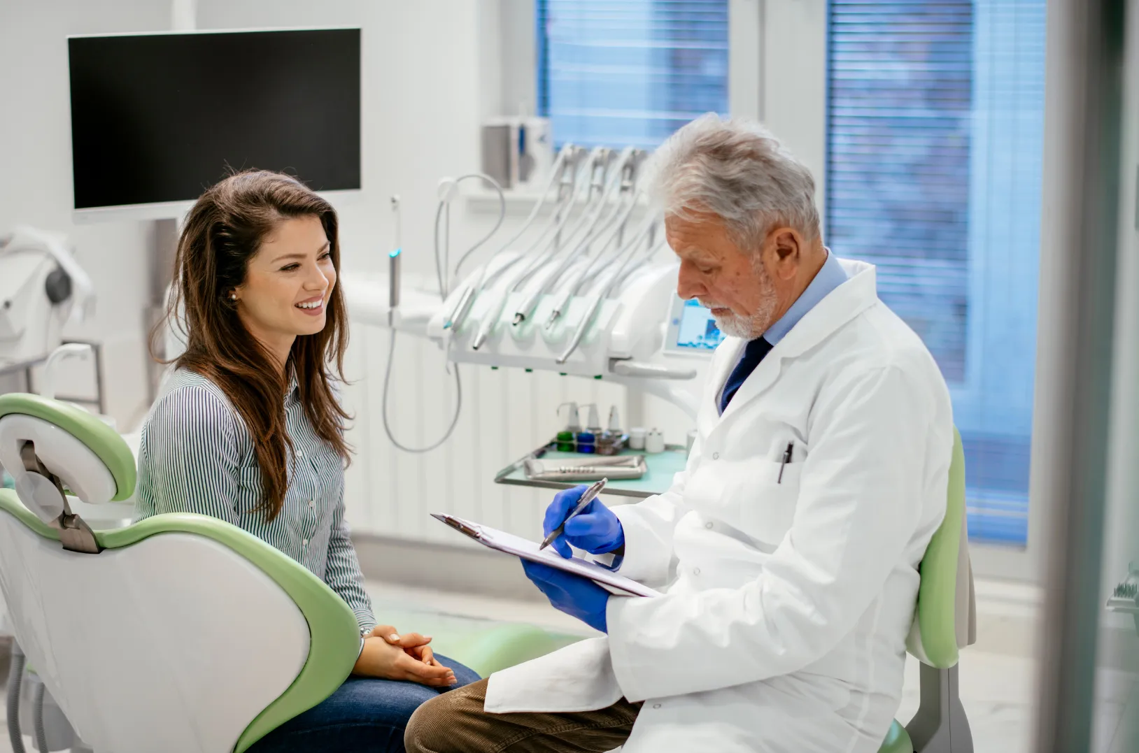 Dentist in white coat and blue gloves writing on clipboard while talking to a smiling female patient seated in a dental chair.