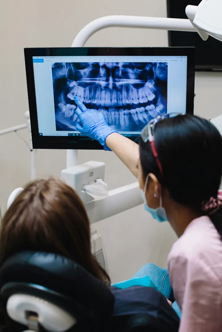 Dentist wearing gloves and a mask pointing to a dental X-ray on a screen while explaining to a seated patient.