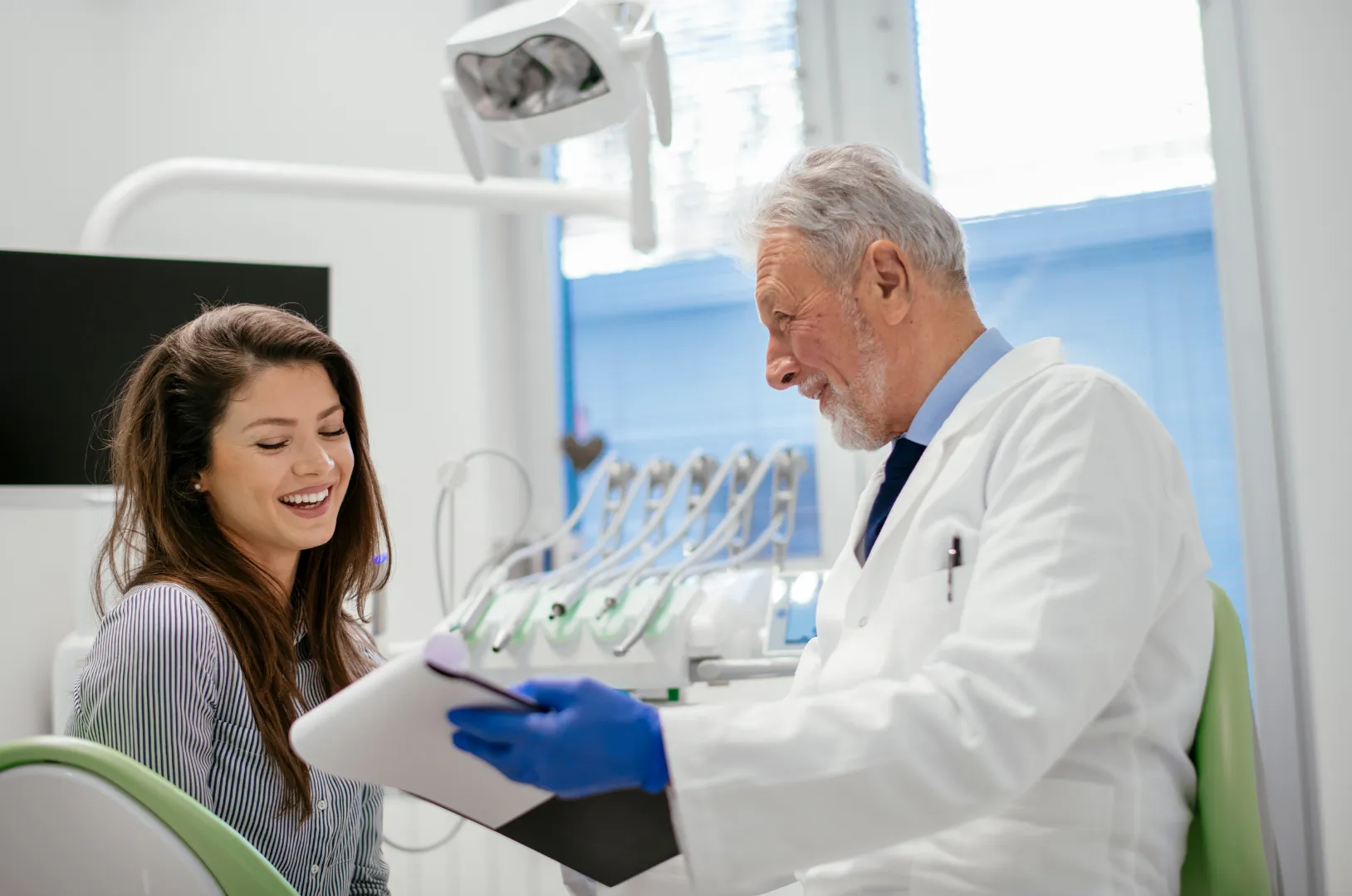 Smiling female patient talking with a male dentist wearing white coat and blue gloves in a dental clinic.
