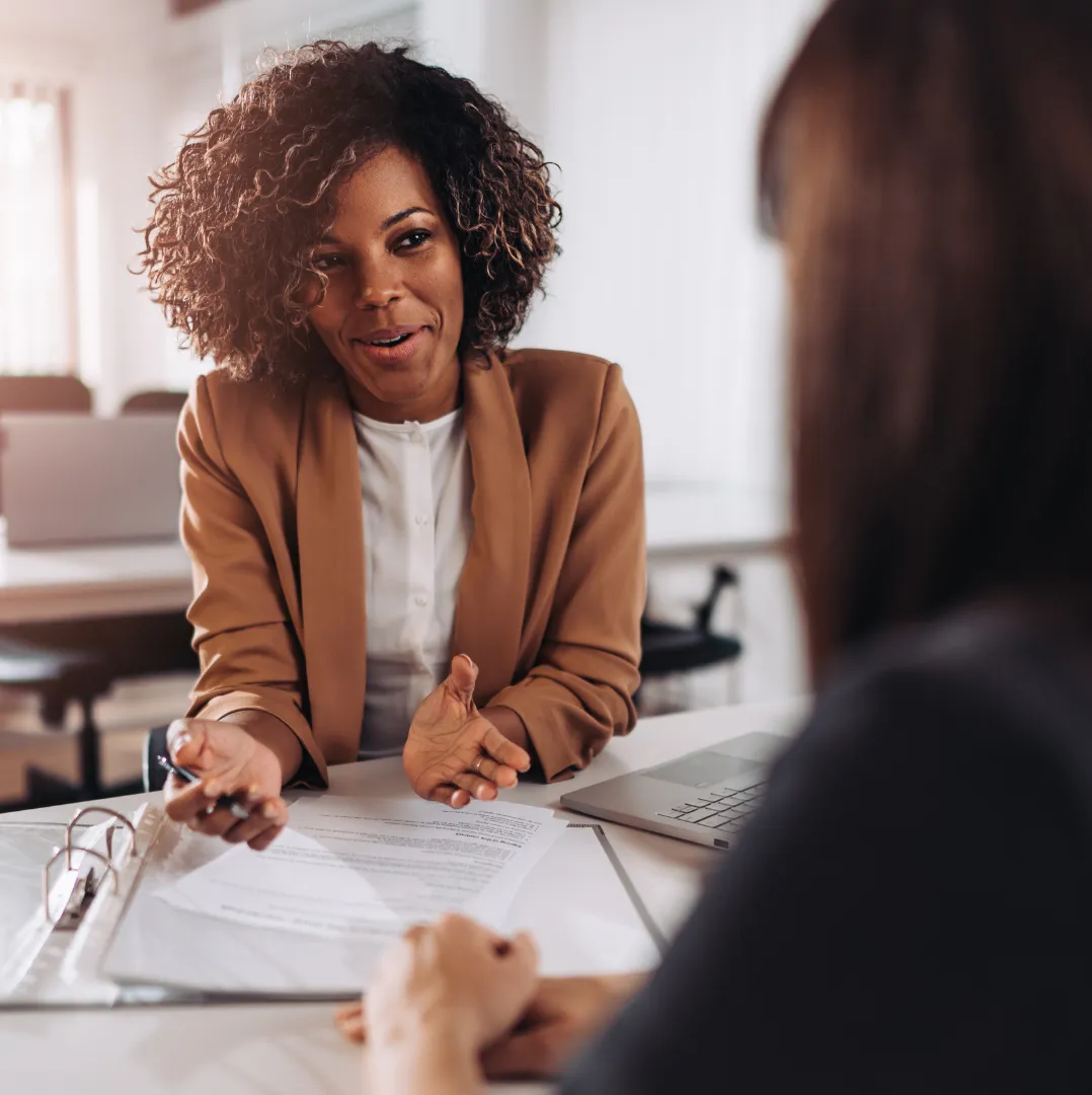 Woman in brown blazer talking and gesturing during a meeting with another person at a desk.