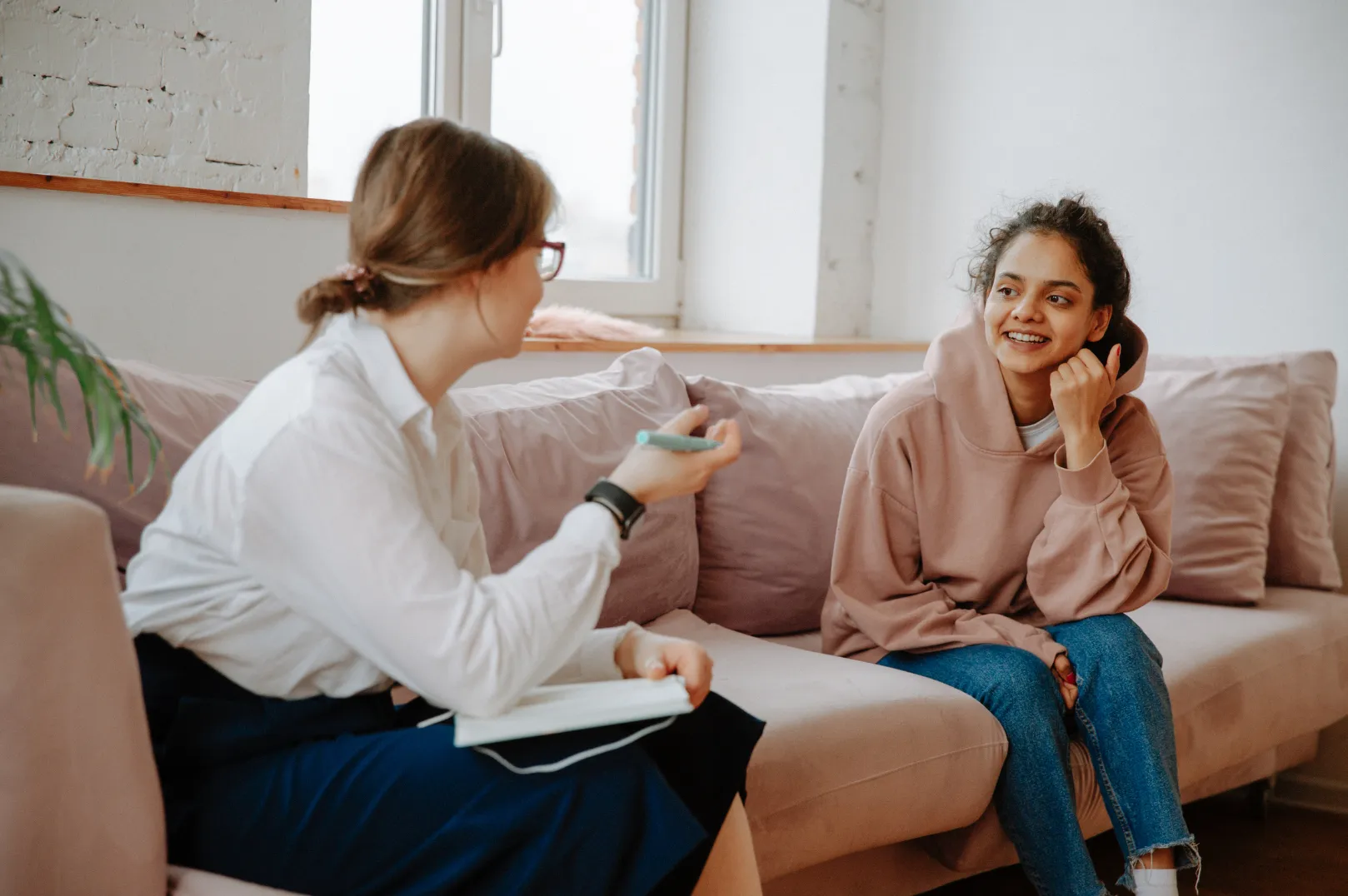 Two women sitting on a beige couch, one taking notes and talking, the other listening and smiling.