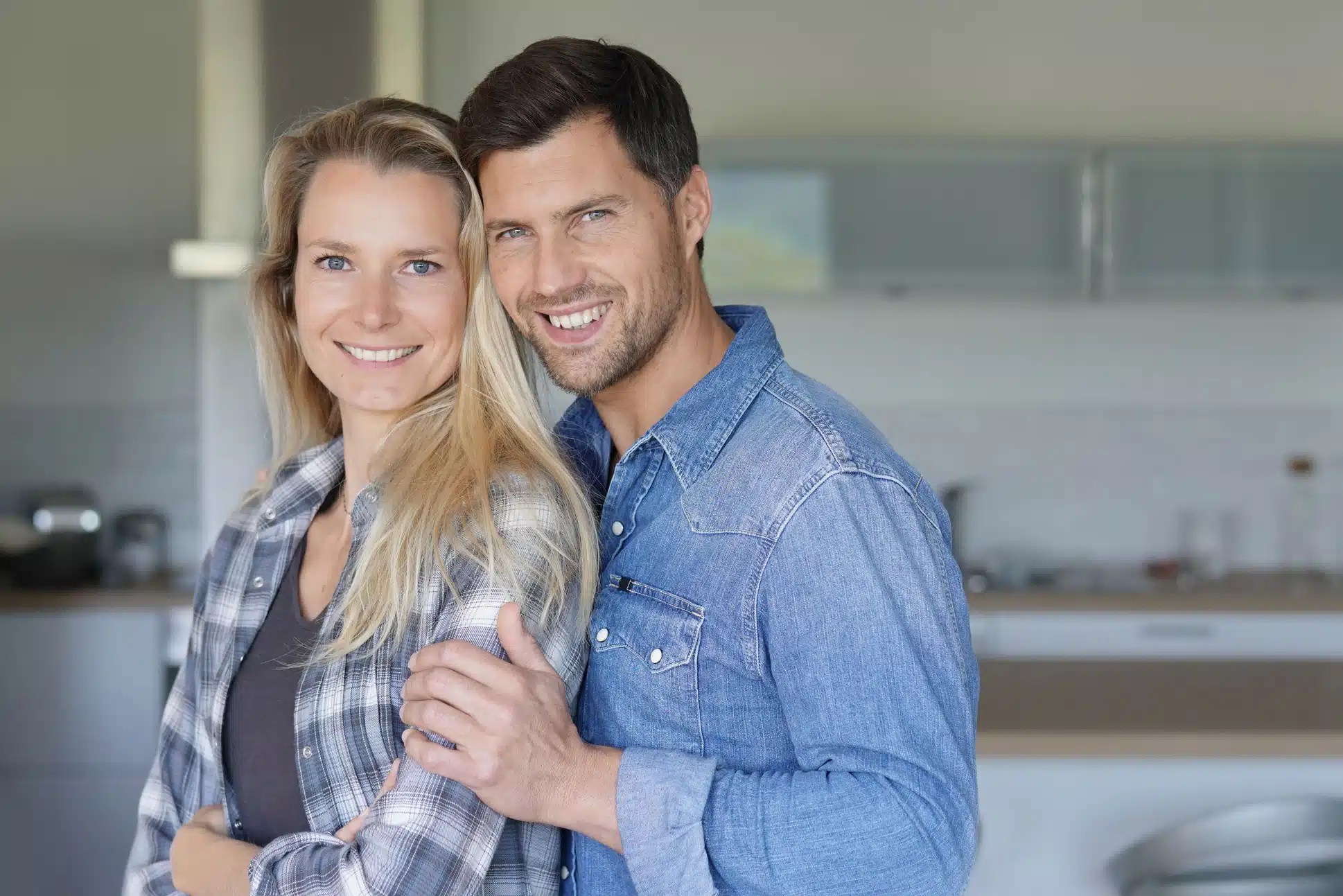 Smiling couple standing close together in a modern kitchen, man in denim shirt holding woman's arm.