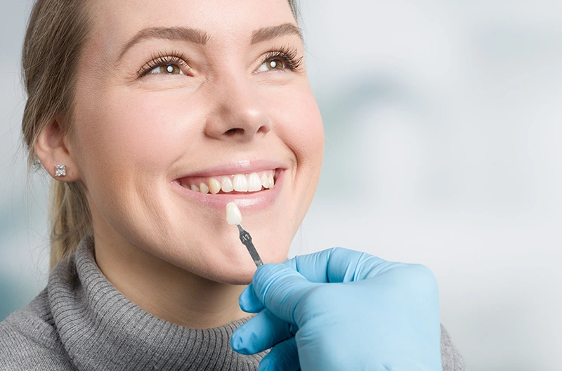 Dentist wearing blue gloves comparing a dental veneer to a smiling woman's upper front teeth.