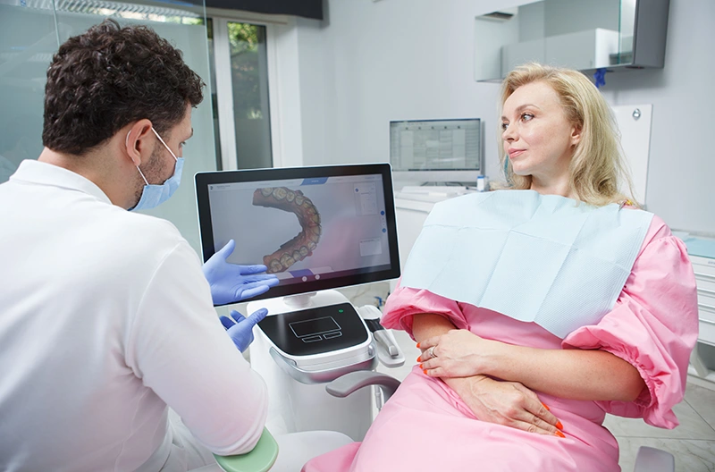 Dentist wearing mask and gloves showing a dental 3D scan on a monitor to a woman patient in a pink gown sitting in a dental chair.