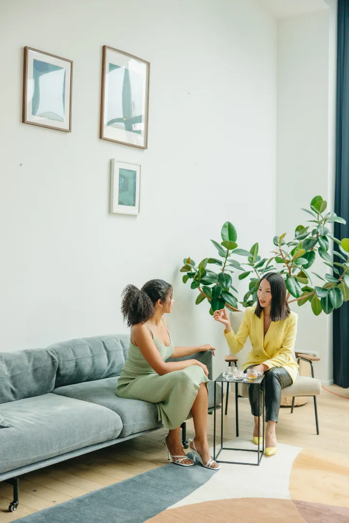 Two women having a conversation in a modern living room; one sitting on a gray sofa wearing a green dress, the other on a chair wearing a yellow blazer and black pants.