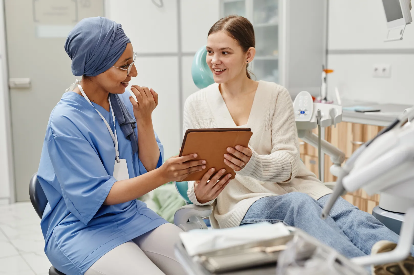 Smiling female healthcare professional in blue scrubs shows tablet to female patient sitting in a dental chair.