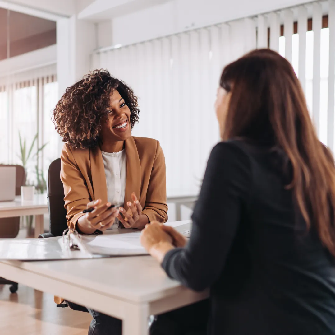 Smiling woman in a brown blazer speaking with another woman across a desk in an office setting.