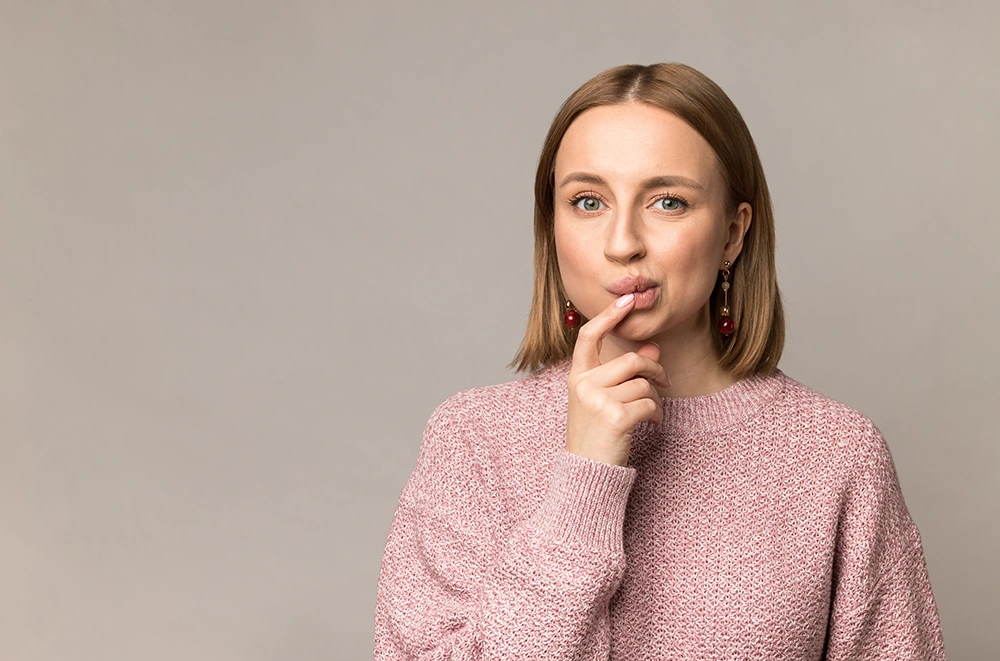 Woman with light brown hair wearing a pink sweater, touching her lips with a thoughtful expression.