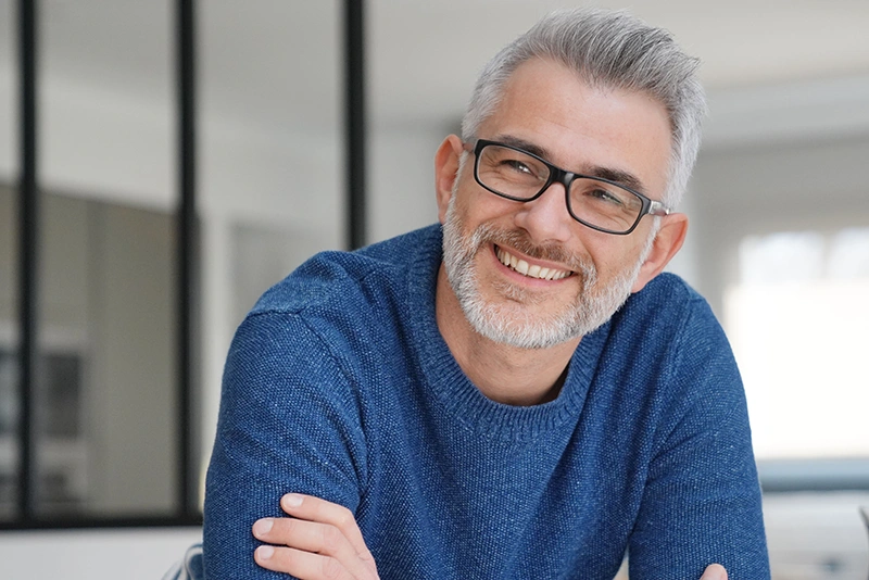 Middle-aged man with gray hair and beard wearing glasses and a blue sweater, smiling indoors.