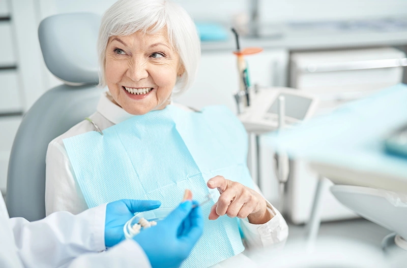 Smiling elderly woman sitting in dental chair wearing a blue bib and interacting with a dentist holding dentures.