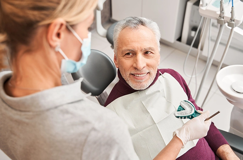 Smiling elderly man sitting in a dental chair while a masked dental professional holds a dental impression tray.