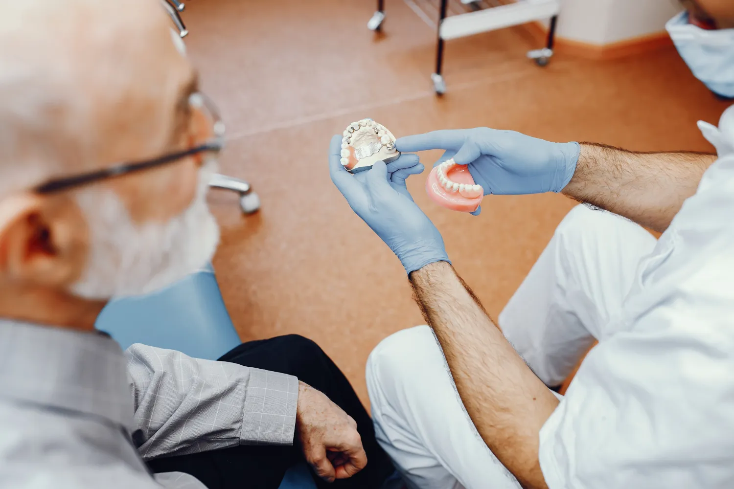 Dentist wearing blue gloves showing a dental mold and dentures to an elderly patient.