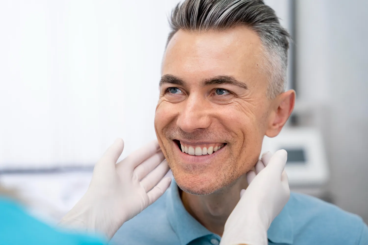 Smiling middle-aged man with gray hair in a dental or medical checkup, being examined by a gloved professional.