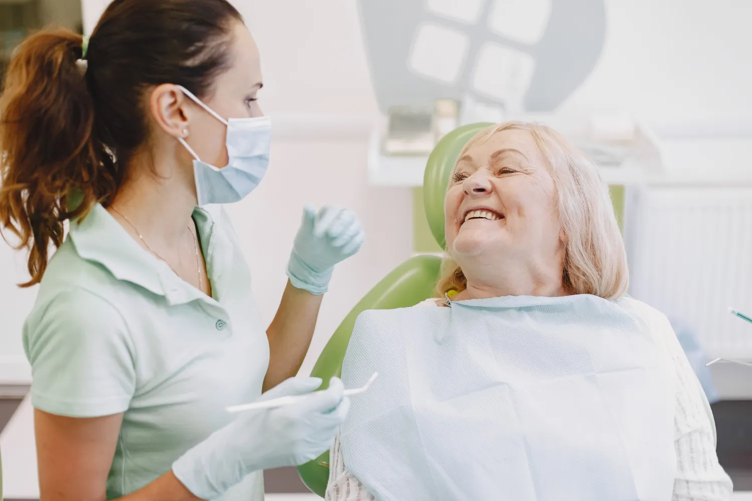 Dentist wearing mask and gloves talking to a smiling elderly female patient seated in a dental chair.