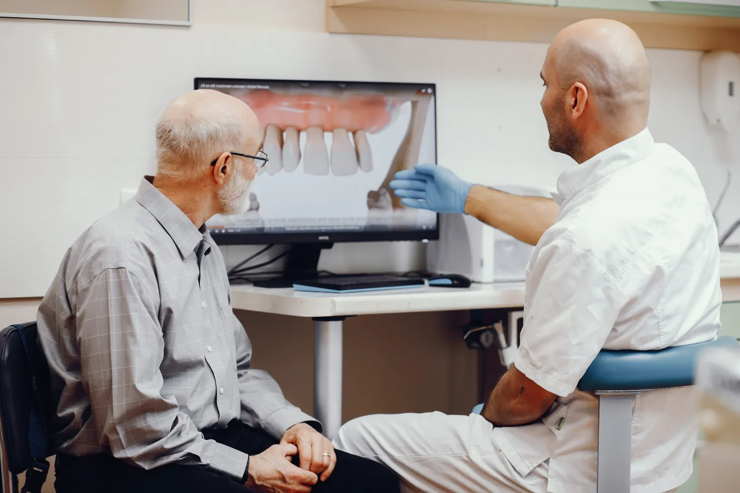 Dentist in white coat explaining dental treatment plan on computer screen to elderly male patient.