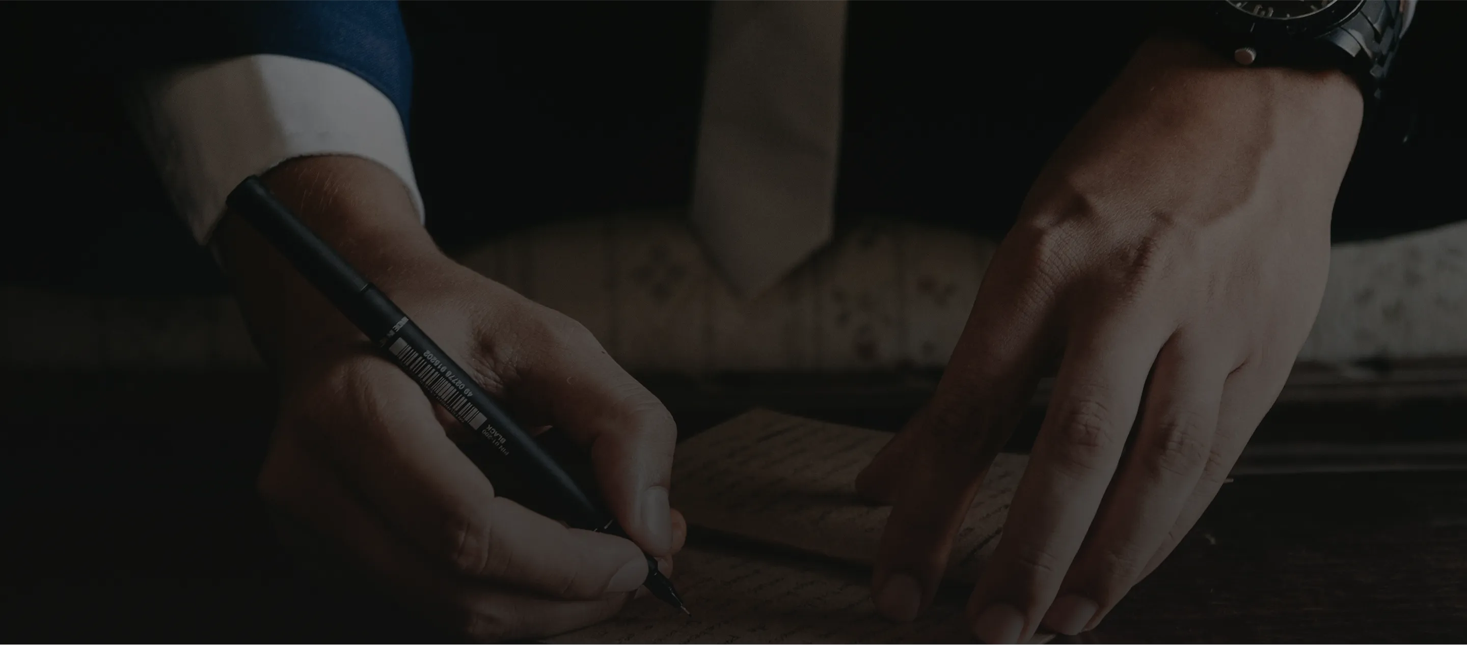 Person in a suit writing with a black pen on a piece of paper on a dark wooden desk.