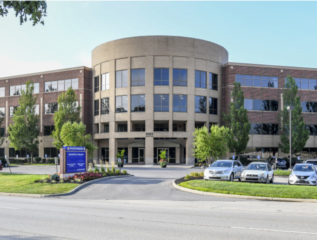 Three-story brick and concrete office building with a semicircular front entrance, surrounded by trees, parked cars, and landscaping.