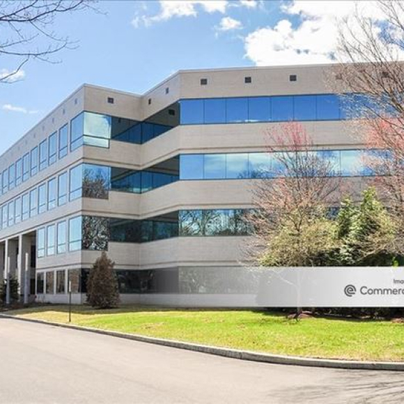 Modern four-story office building with reflective blue windows, surrounded by trees and a lawn under a partly cloudy sky.
