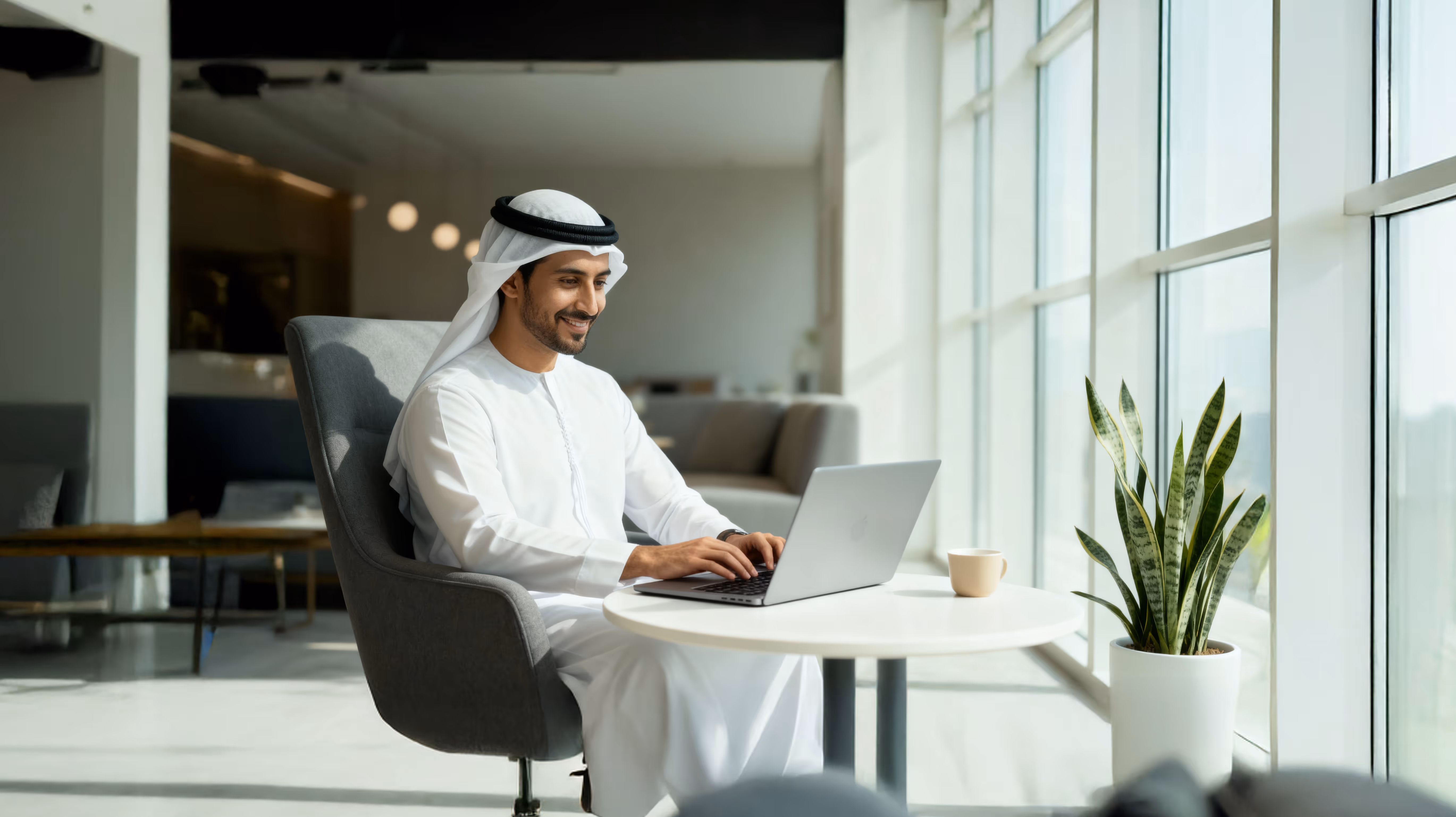 Smiling man in traditional Middle Eastern attire working on a laptop at a round table by large windows.