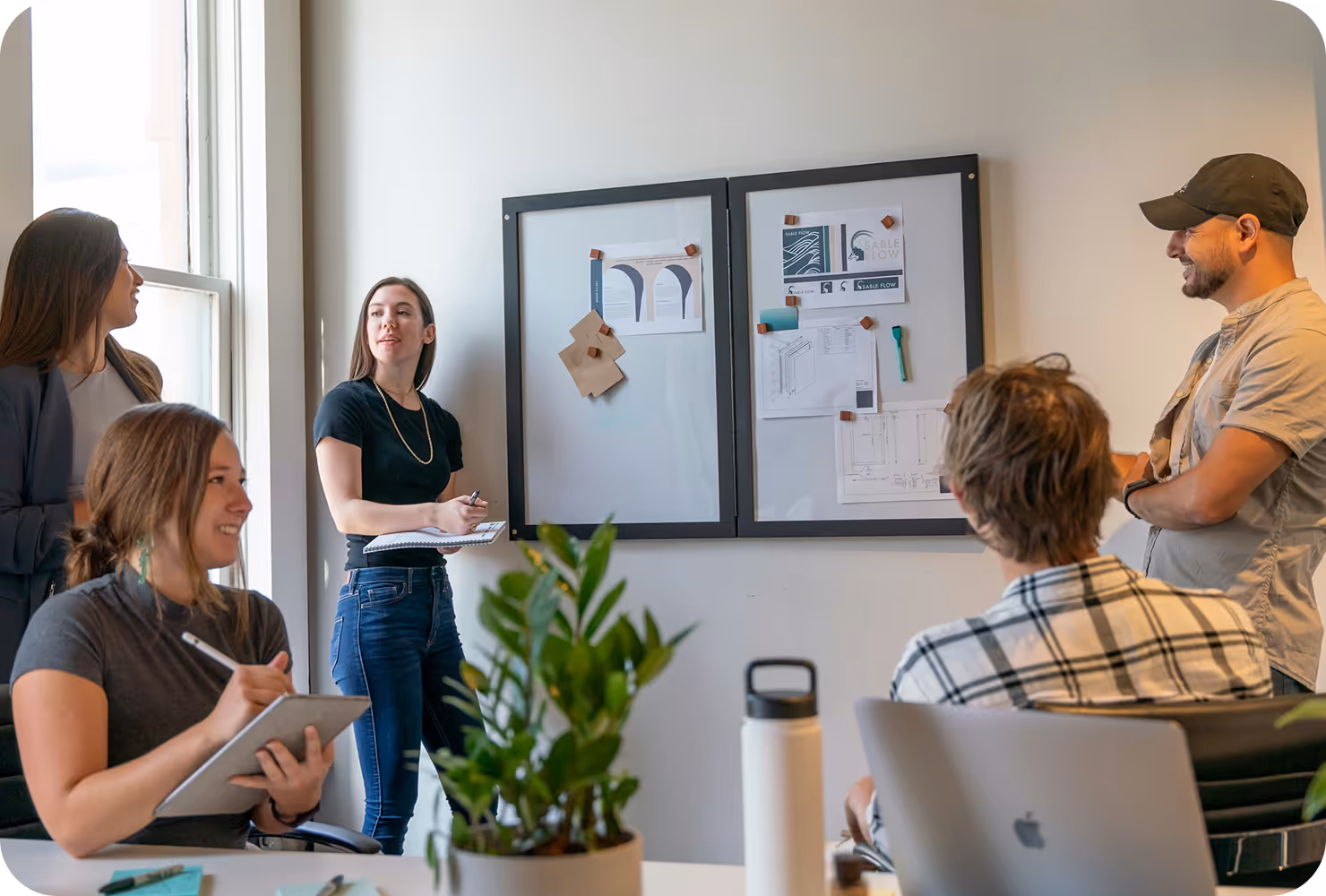 Five colleagues in a modern office engaged in a discussion around a whiteboard with pinned documents and sketches.
