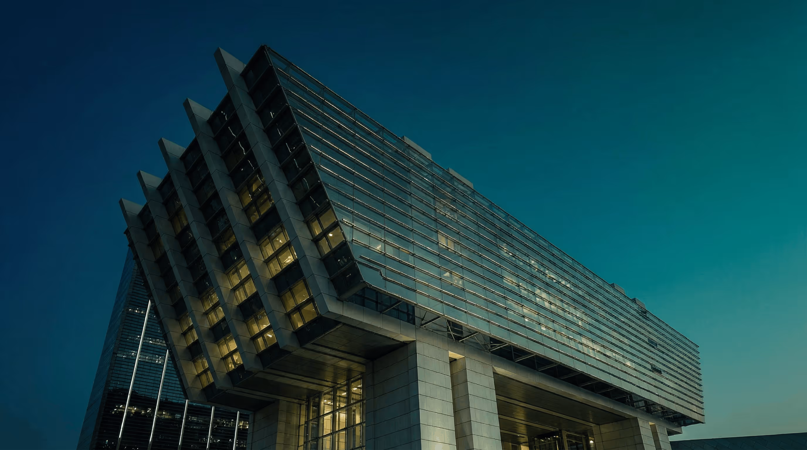 Modern glass office building at dusk with illuminated interior and a dark blue sky.