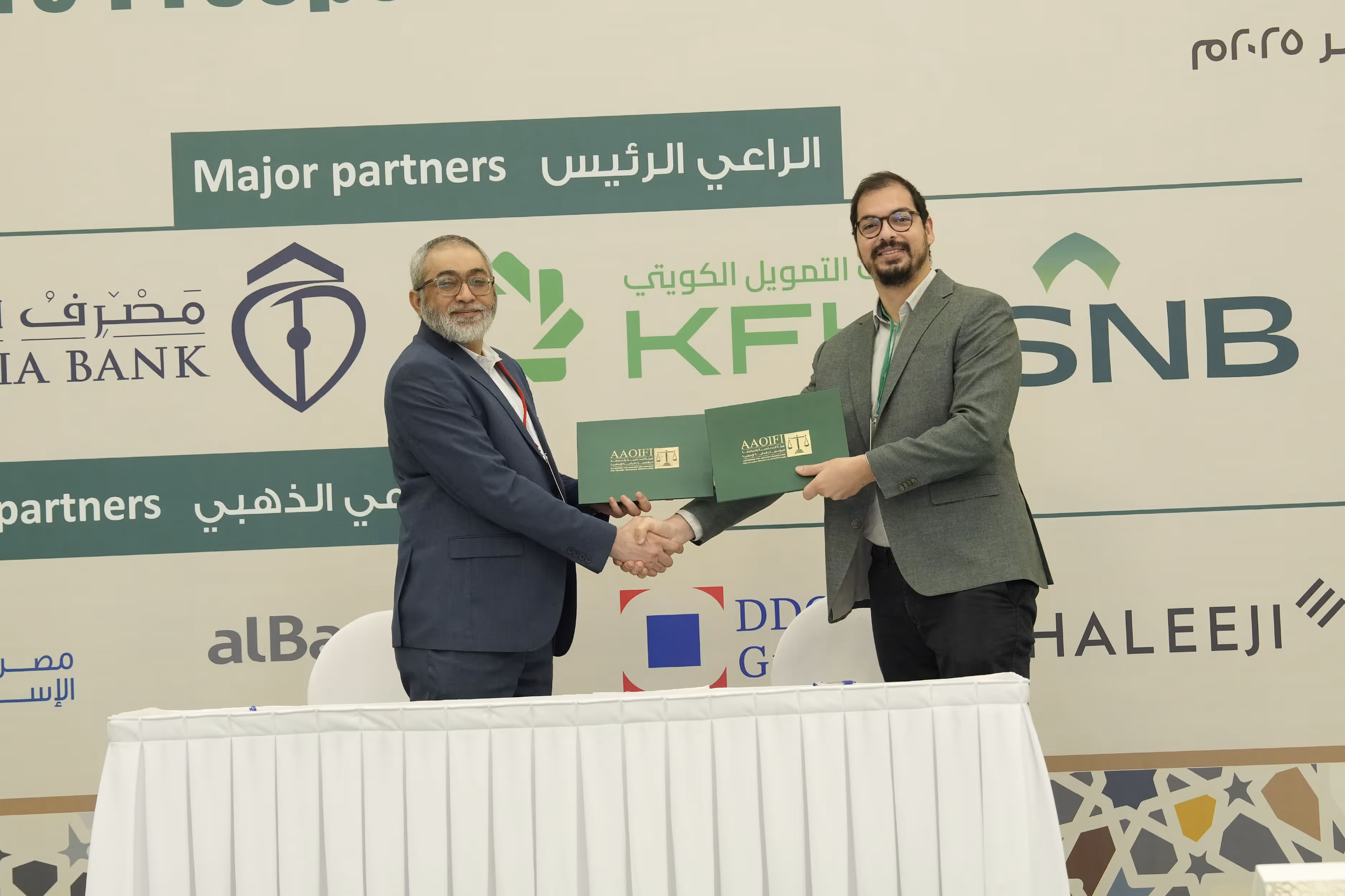 Two men in suits shaking hands and holding folders during a formal partnership signing event in front of a backdrop displaying major partner logos.