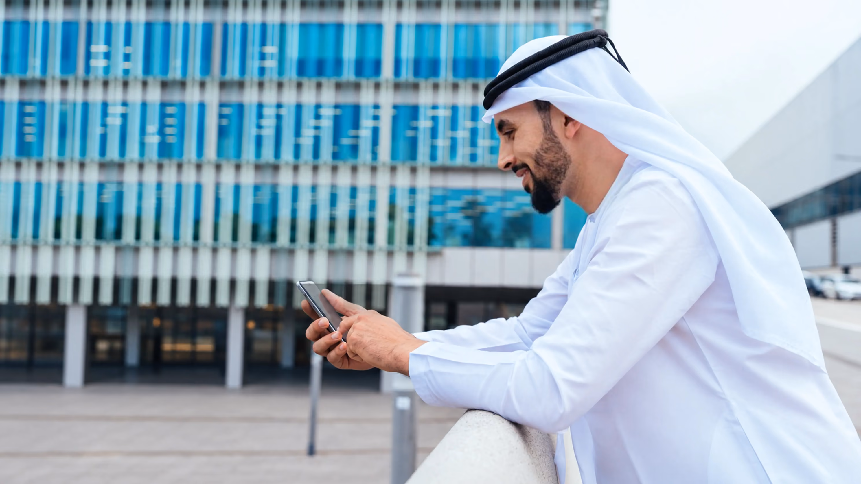 Man wearing traditional Middle Eastern white clothing and headscarf leaning on a railing outdoors while smiling and using a smartphone with a modern glass building in the background.