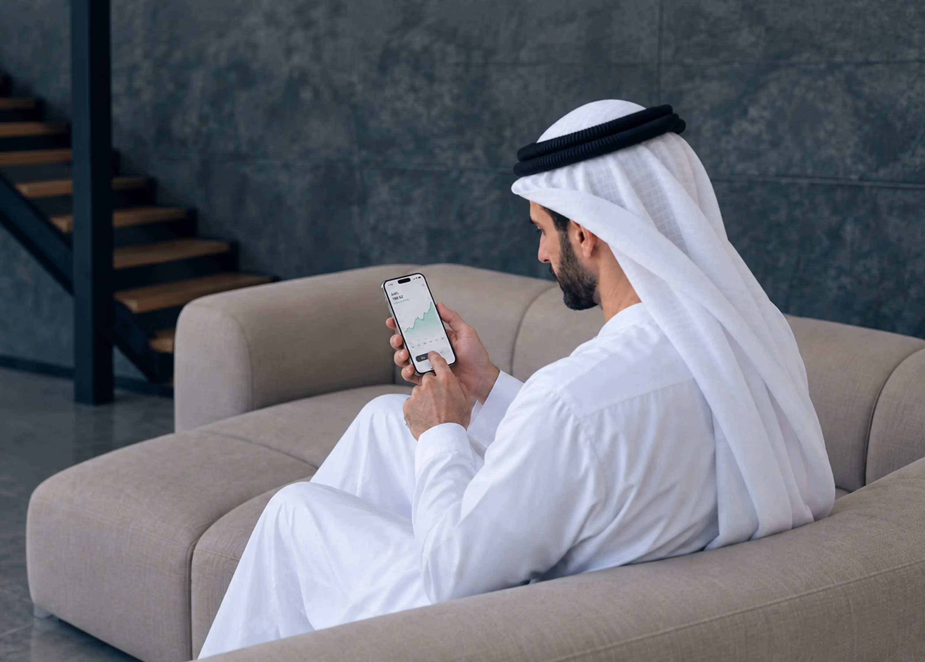 Man wearing traditional Middle Eastern attire sitting on a beige sofa looking at a smartphone displaying a green upward trending graph.
